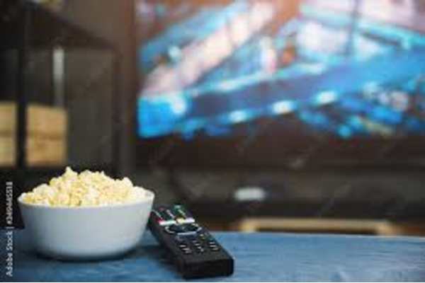 A bowl of popcorn and a remote control on a table in front of a blurred television screen.